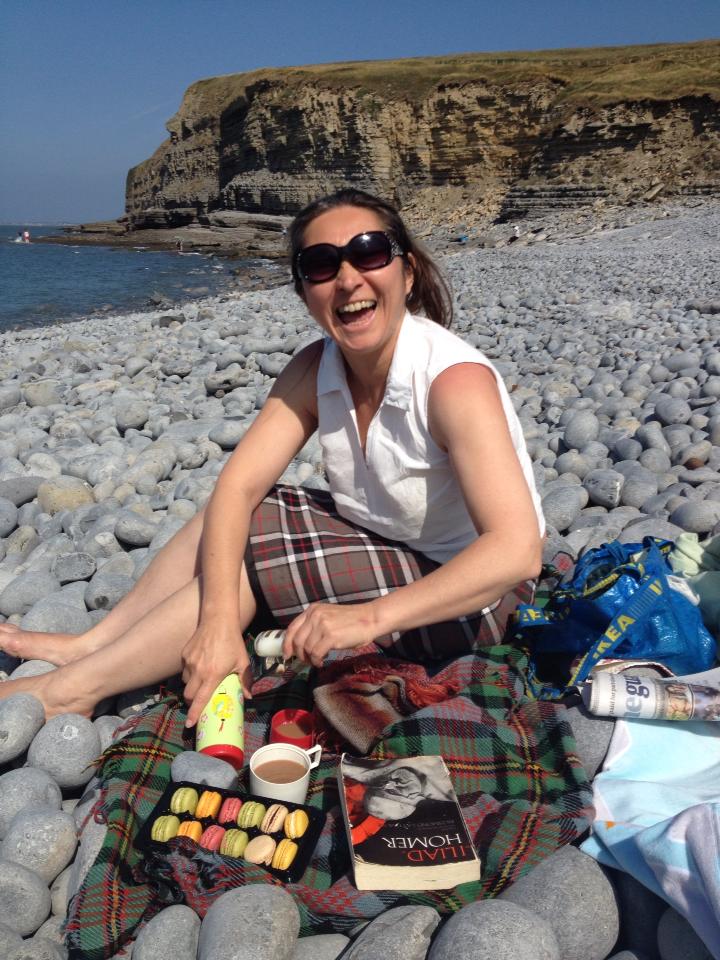 Anita Pilgrim sitting on a pebbly beach in the sunshine, laughing