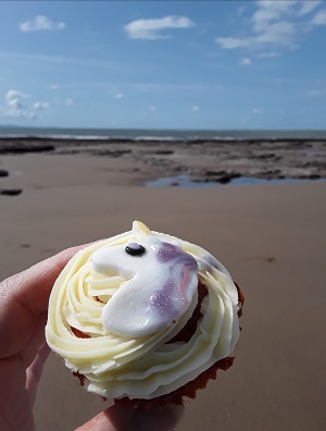 Fingers holding a unicorn cupcake with a beach in the background