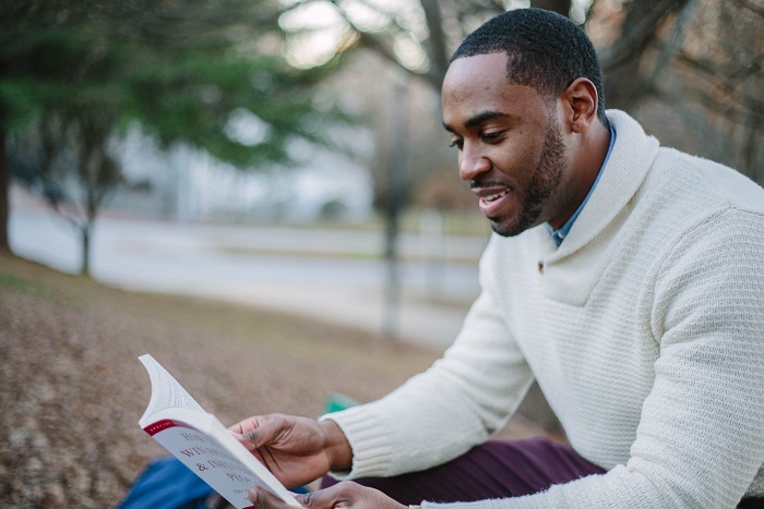 A young bearded black man reading a book outdoors near a lake