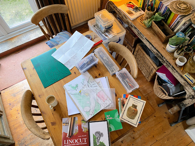 A dining table covered in printmaking materials, sketches, vinyl, paper and tools.