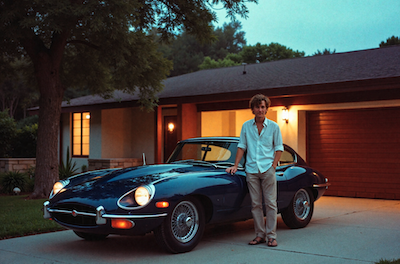 A young man stands next to a blue E Type Jag outside a California house