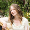 Head and shoulders of a woman in a white dress in front of a hedge holding gold sticks